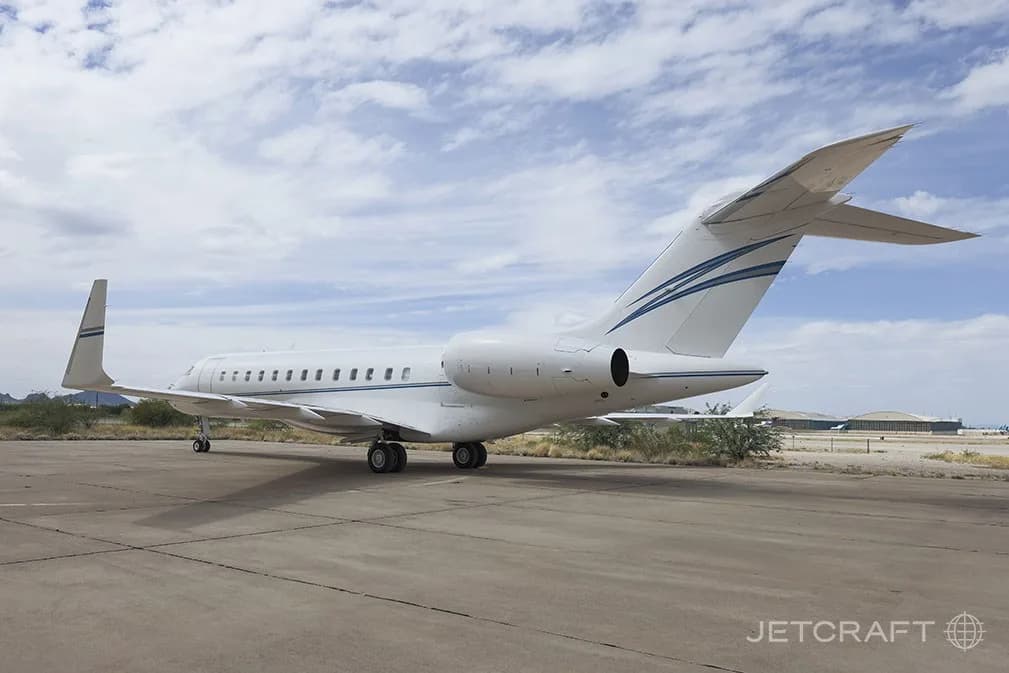 A rear-quarter view of a white Bombardier Global 6000 business jet with blue and grey stripes on the tail, parked on an airfield.