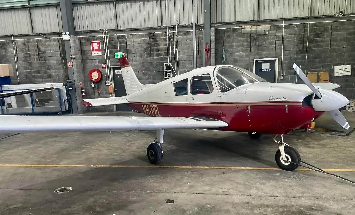 A red and white Piper Cherokee 180 aircraft, registration VH-PEL, is parked inside a hangar with a concrete block wall.