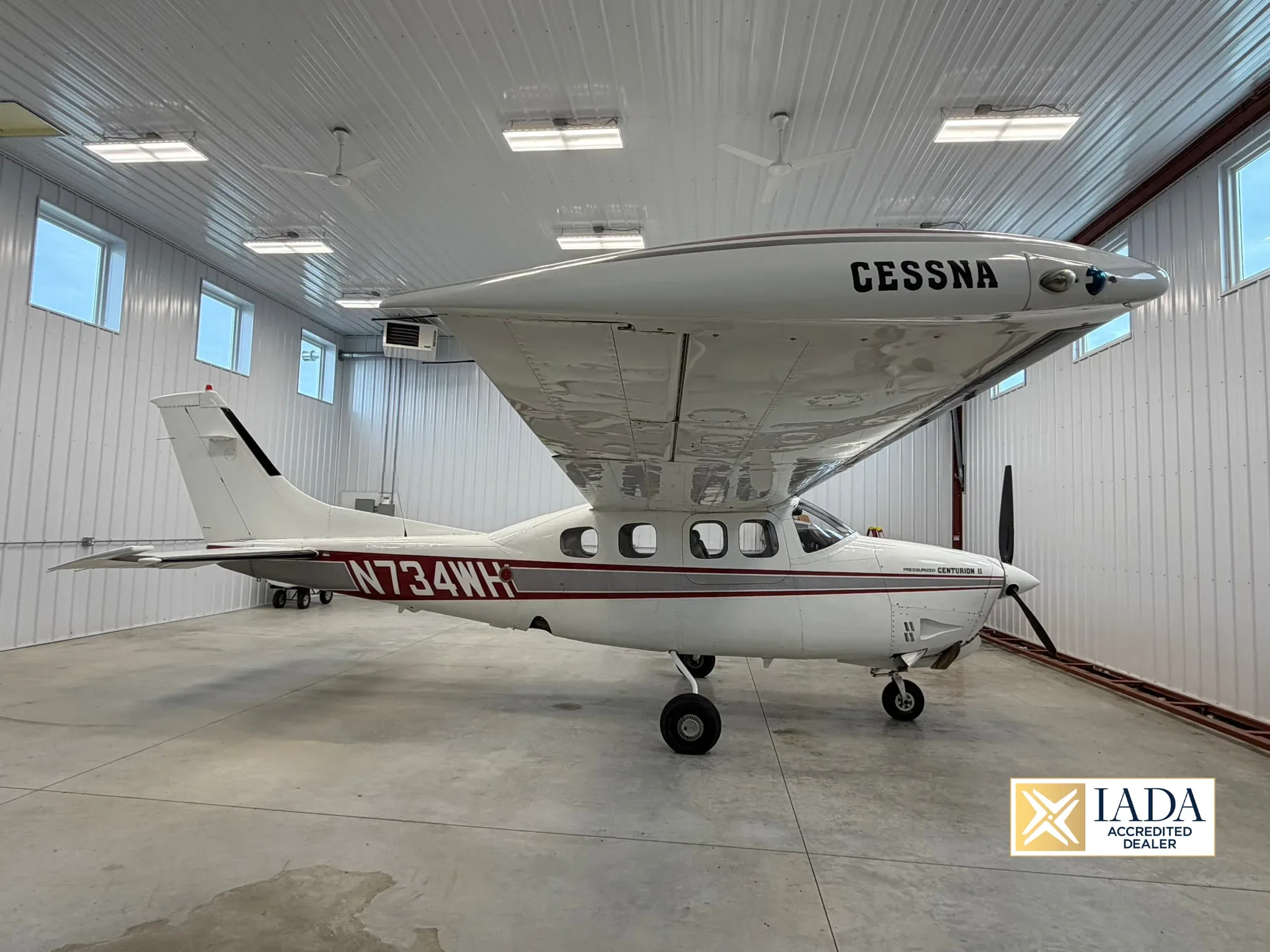 A white Cessna Pressurized Centurion II (N734WH) with red and grey stripes, parked inside a well-lit hangar.