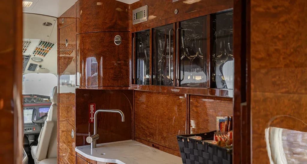 The galley area of a business jet cabin, featuring polished wood cabinetry, a sink, and glassware storage.