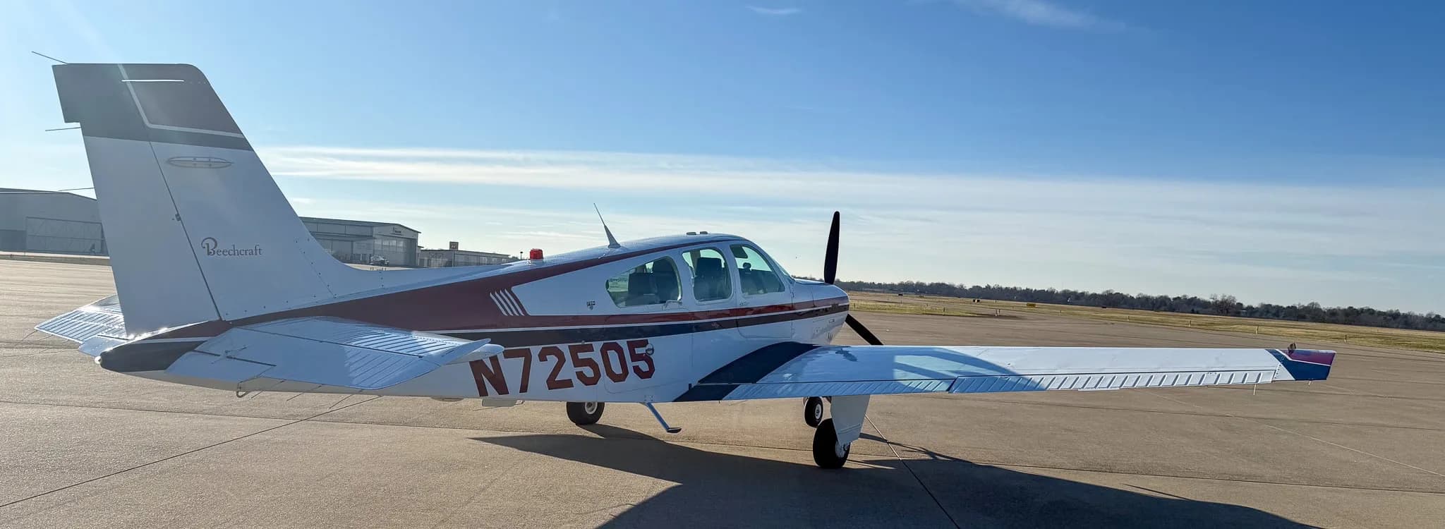 Rear-left view of a white Beechcraft Bonanza with red and blue stripes, showing registration N72505, on an airfield.