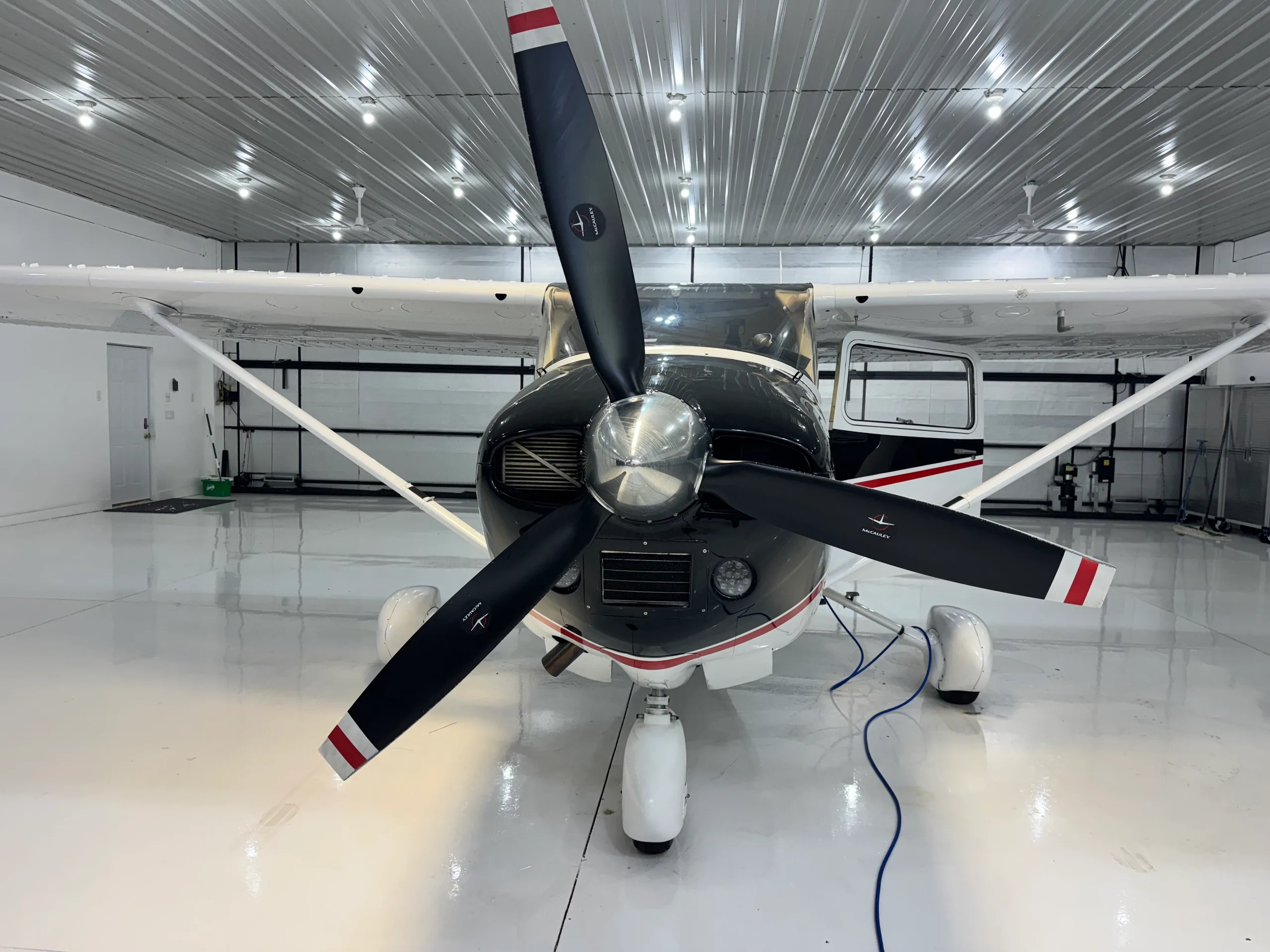 A head-on view of a black and white Cessna 182XP aircraft with a three-bladed propeller, parked in a hangar.
