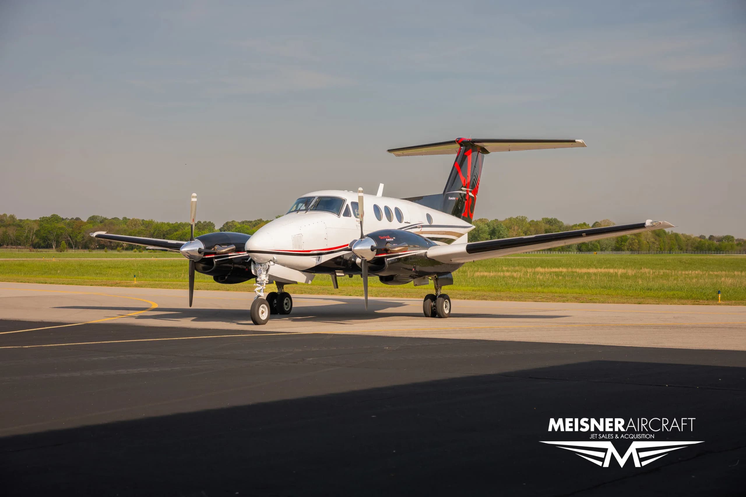 A white and black Beechcraft King Air with red tail accents parked on an airfield, viewed from the front-right.