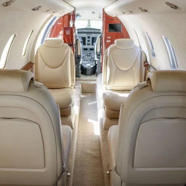 View from the rear of the private jet cabin looking forward towards the cockpit, showing beige seats and a narrow aisle.