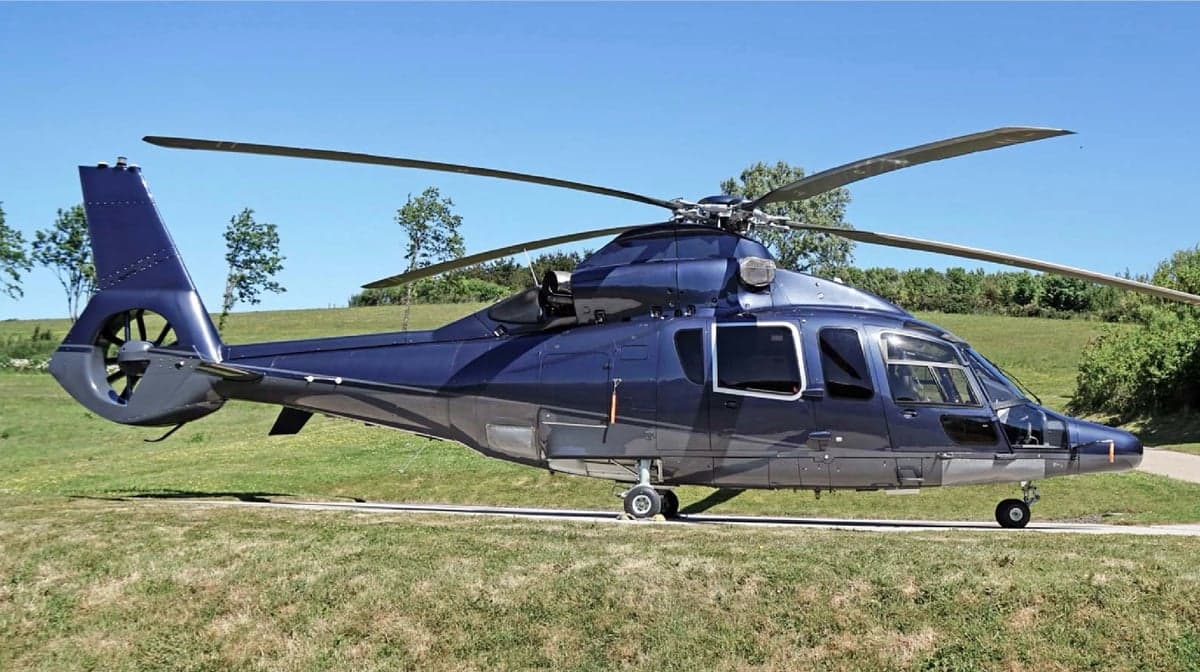 A dark blue helicopter with a grey underside sits on a grassy field on a sunny day.