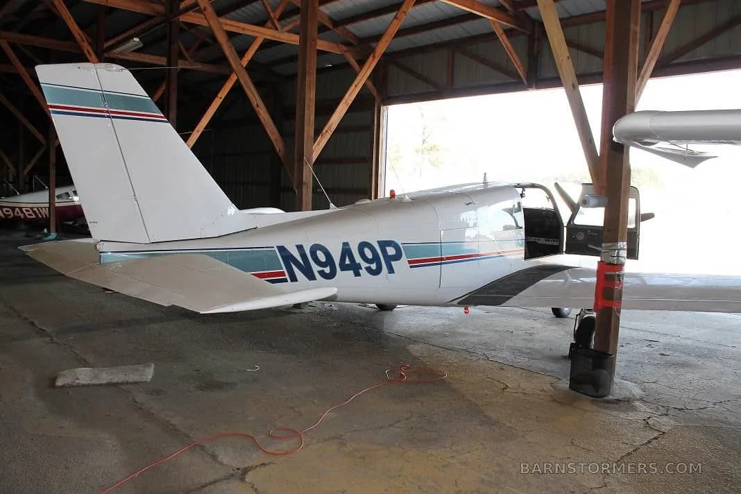 The rear view of a white single-engine propeller aircraft with blue and red stripes, registration N949P, parked in a hangar with its door open.