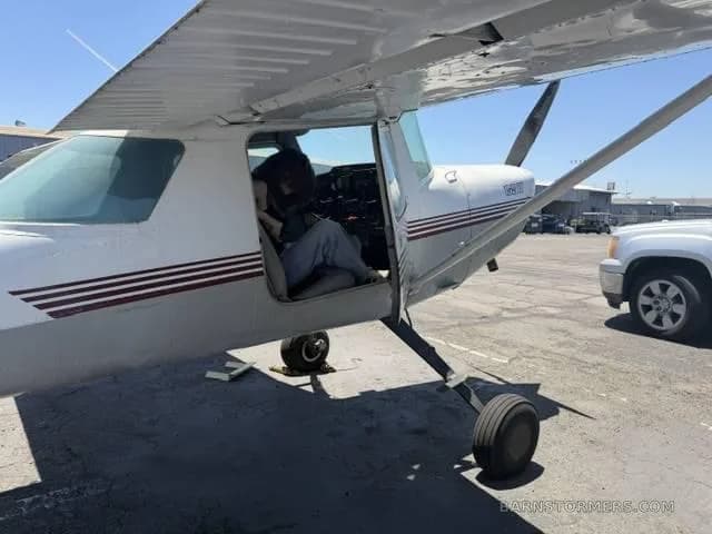 A white and grey single-engine propeller aircraft with its door open, showing a person sitting inside the cockpit, parked on an asphalt ramp.