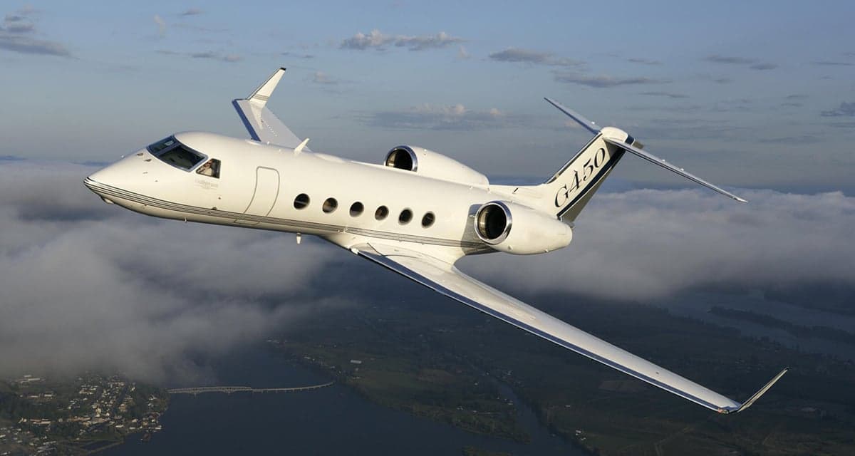 A white Gulfstream G450 business jet with black accents flying over a river and land, seen from below and slightly ahead.