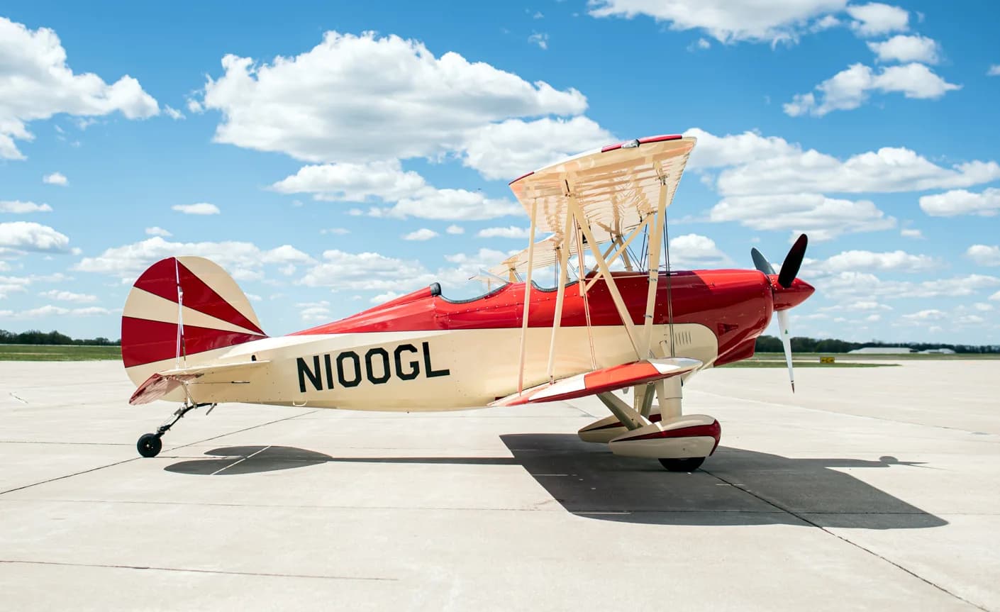 A red and white biplane with tail number N100GL is parked on the tarmac with a blue sky background.