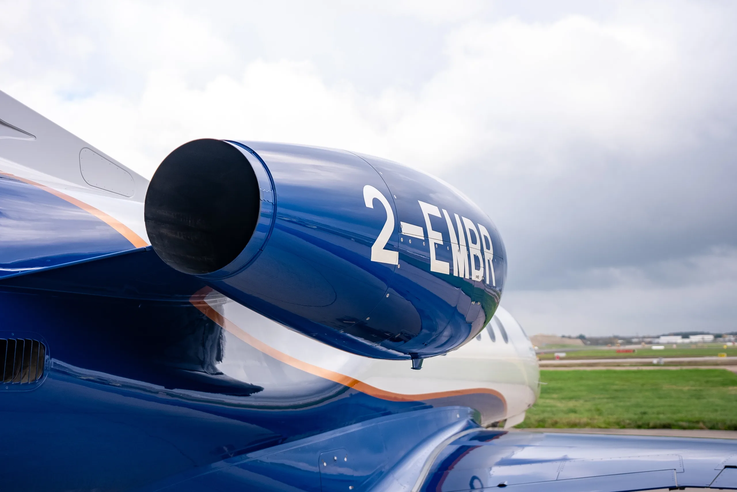 A close-up view of the blue engine nacelle of an Embraer Phenom 300E, showing the registration "2-EMBR".