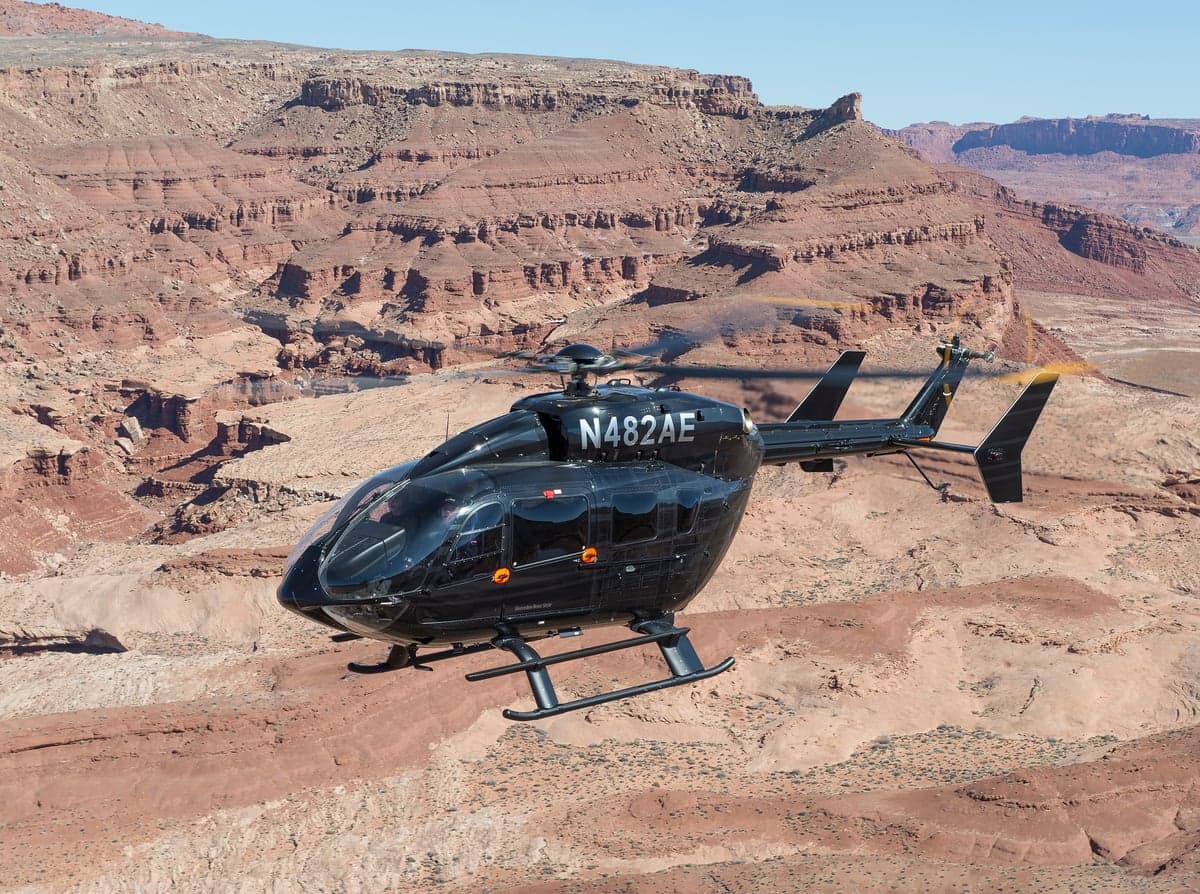 A black Airbus H130 helicopter, registration N482AE, flying over a desert landscape.