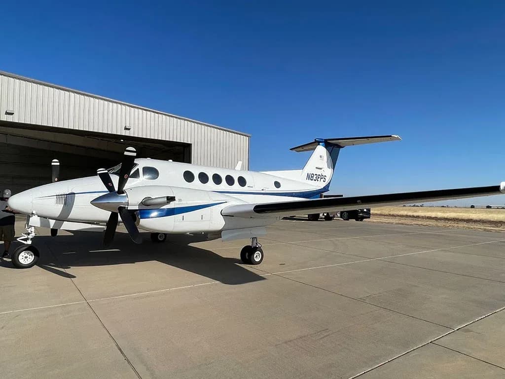 A white and blue twin-engine turboprop aircraft, N838PS, parked on an airfield next to a hangar.