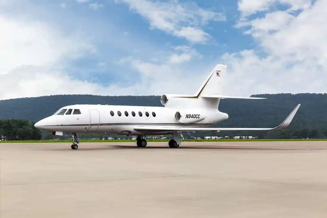 A Falcon jet sits on the tarmac with mountains in the background.