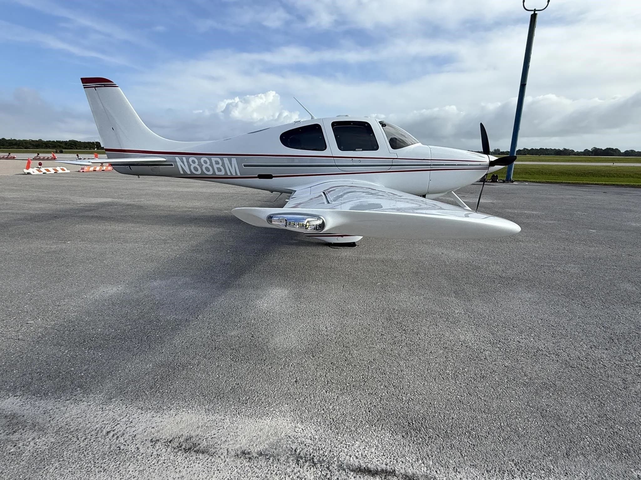 A white Cirrus SR22 aircraft with red and gray stripes, tail number N88BM, parked on an airport tarmac, showing the right wing and landing gear.