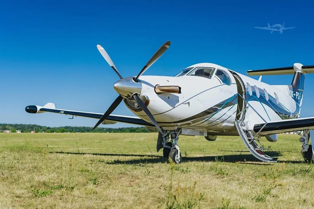 A close-up front view of a white Pilatus PC-12 aircraft with its main cabin door open, parked on a grassy field.