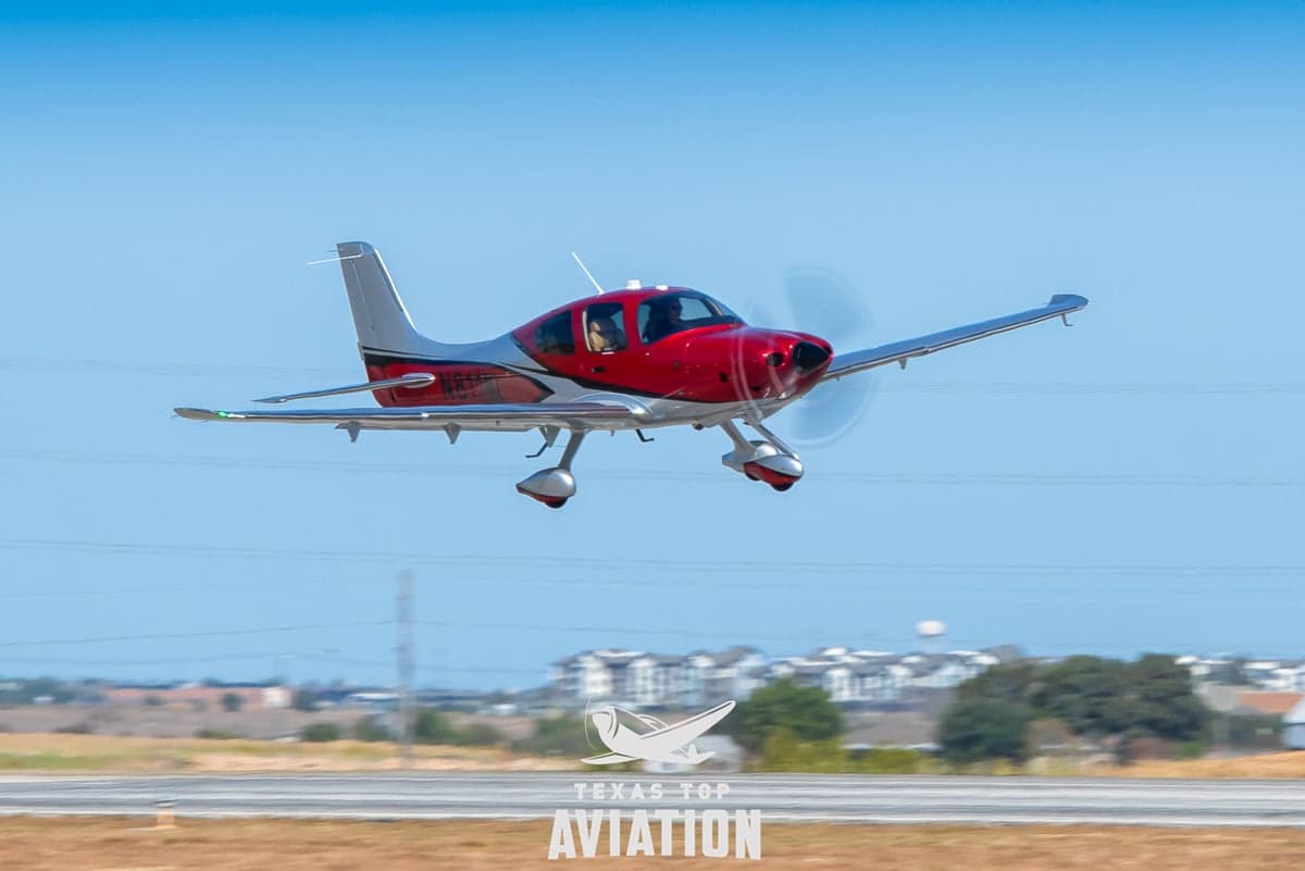 A Cirrus aircraft is taking off with a red and silver paint scheme.