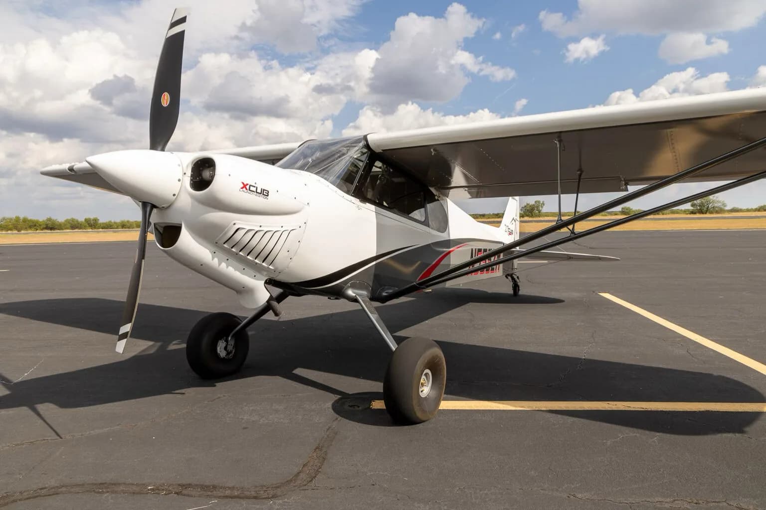 A close-up three-quarter front view of the white and grey X-Cub aircraft N555ZM with red accents, parked on an asphalt runway.