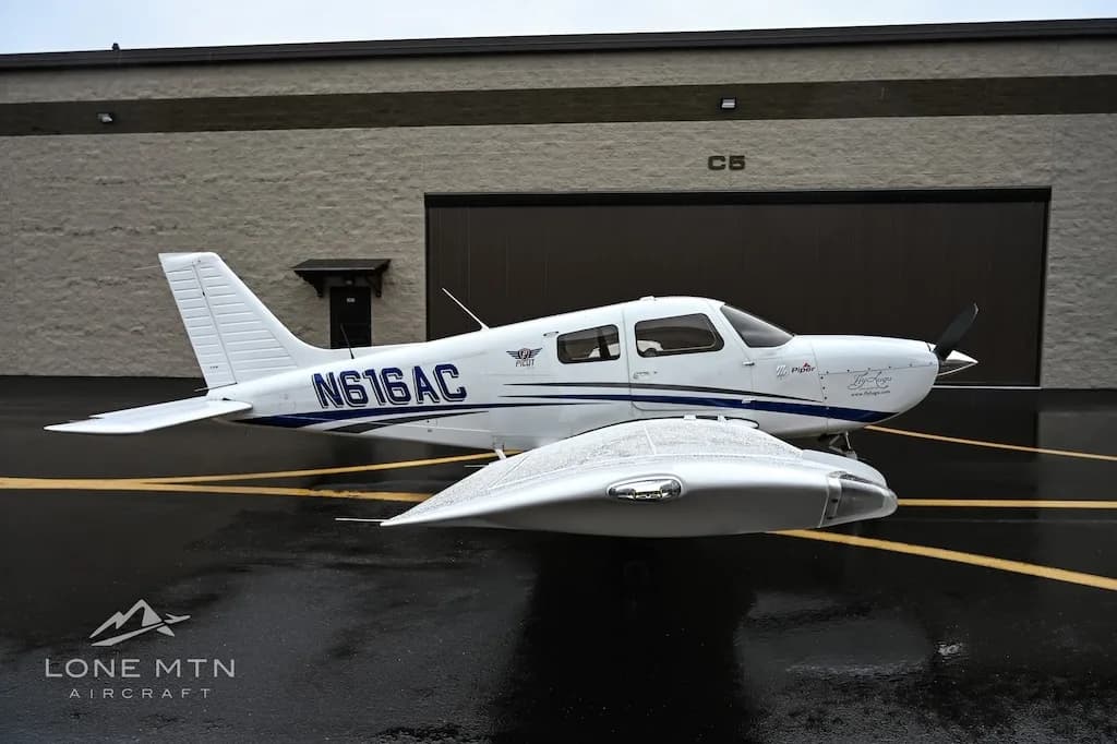 A white single-engine aircraft, N616AC, with blue stripes and wingtip tanks is parked on a wet tarmac.