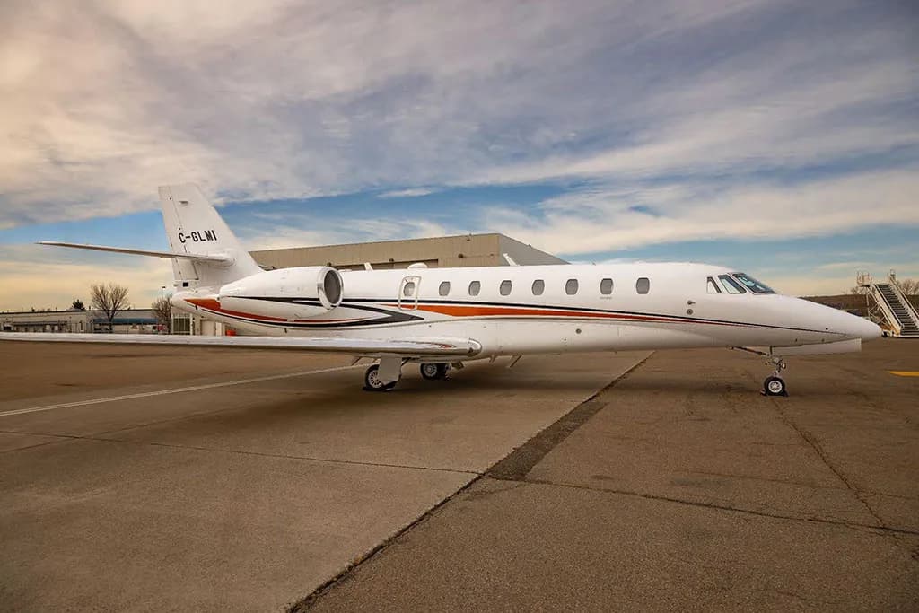 A white business jet with orange and black stripes, parked on an asphalt tarmac with a building and stairs in the background under a partly cloudy sky.