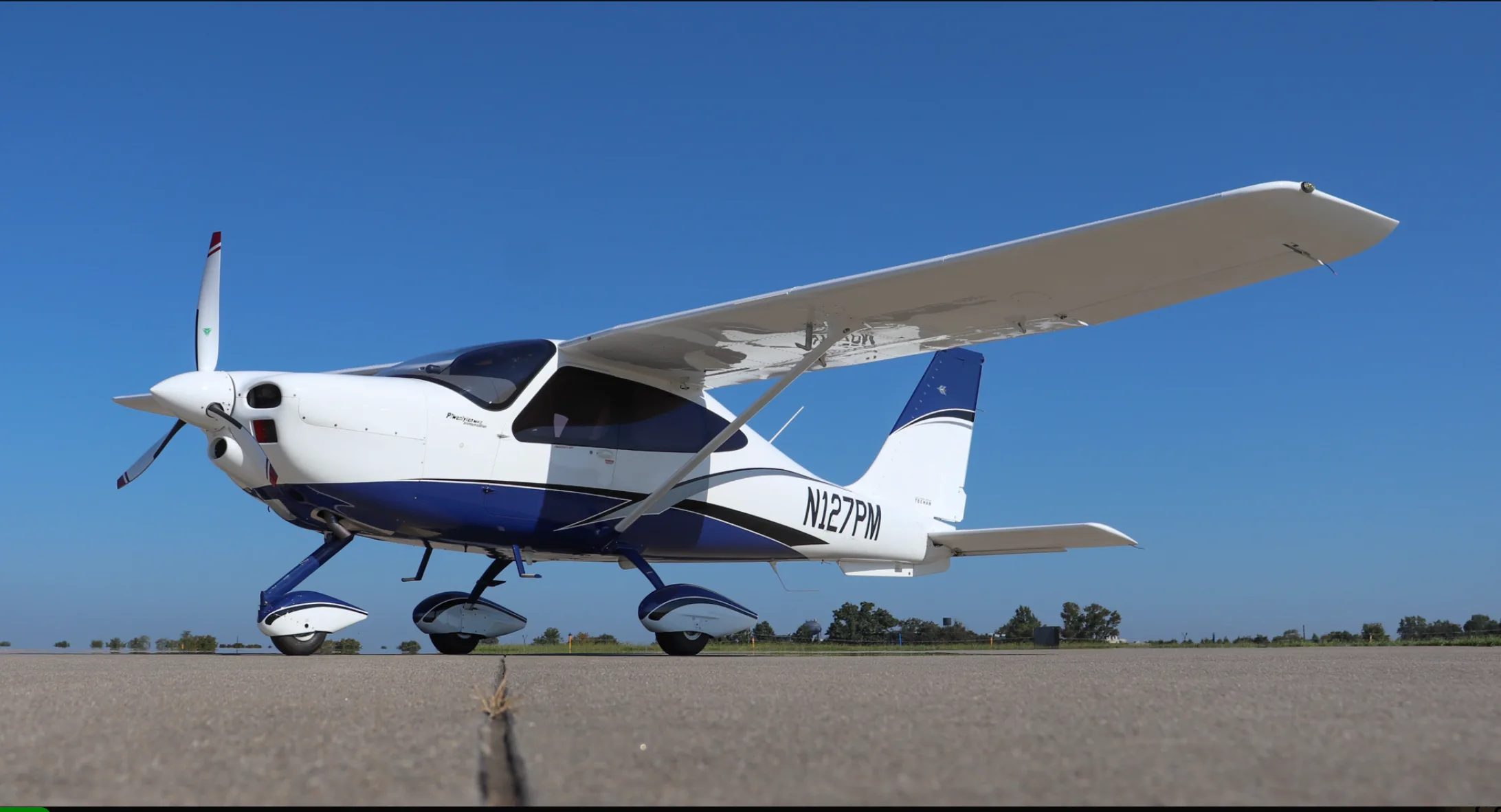 A white and blue Tecnam P2010 aircraft, registration N127PM, parked on an airfield under a clear blue sky.