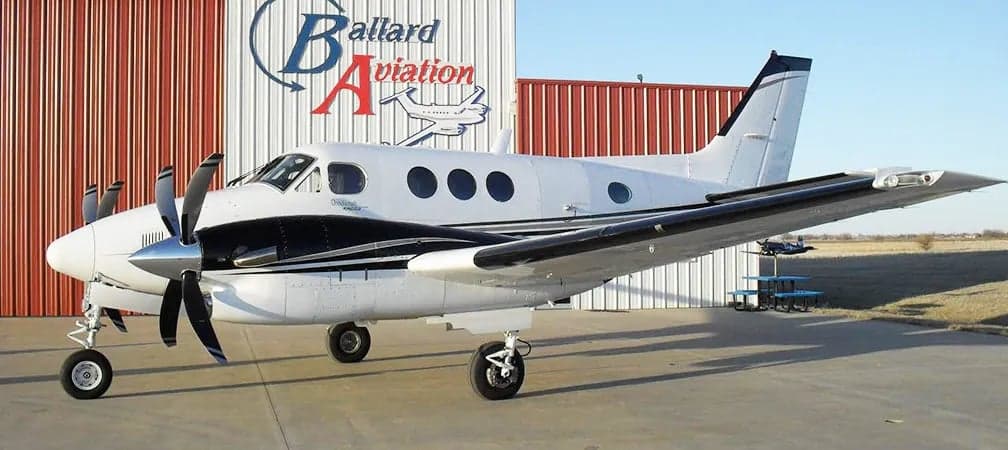 A Beechcraft King Air is parked in front of a hangar with the Ballard Aviation logo.