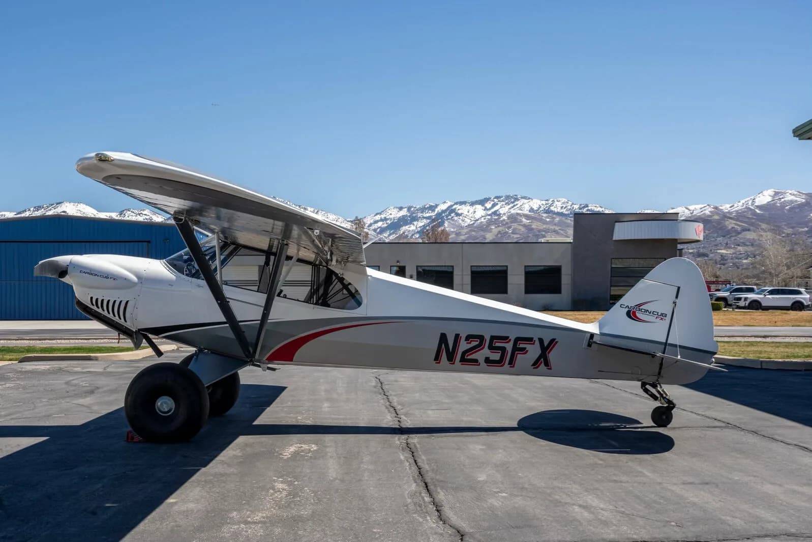 A white and grey Carbon Cub FX-3 aircraft with red accents and registration N25FX parked on an asphalt ramp with snowy mountains in the background.