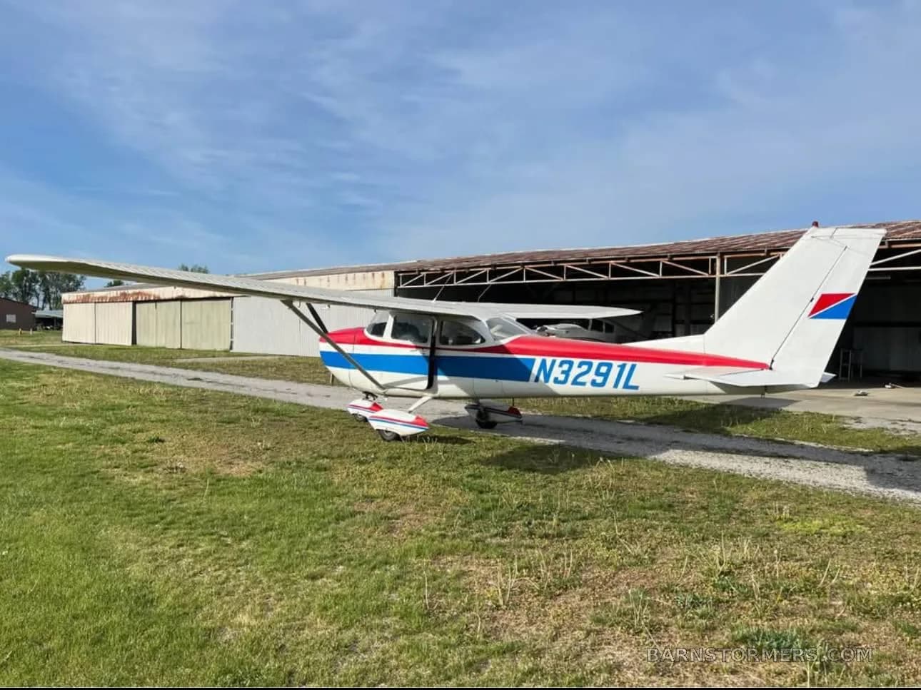 A three-quarter rear view of a white, red, and blue Cessna-style aircraft with registration N3291L parked on a gravel path.