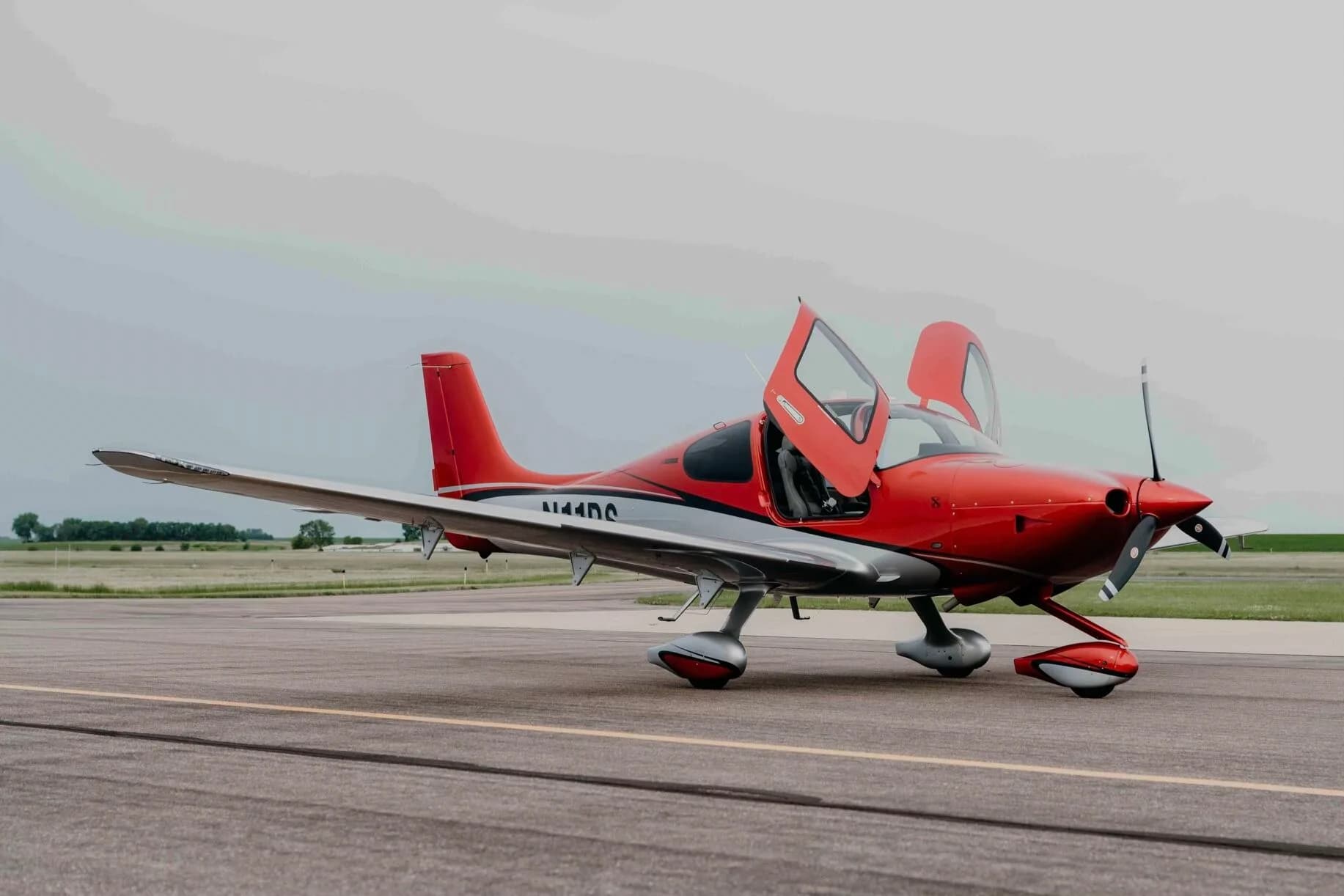 A red and silver Cirrus SR20 Premium aircraft (N11PS) with both doors open, parked on an airfield tarmac.