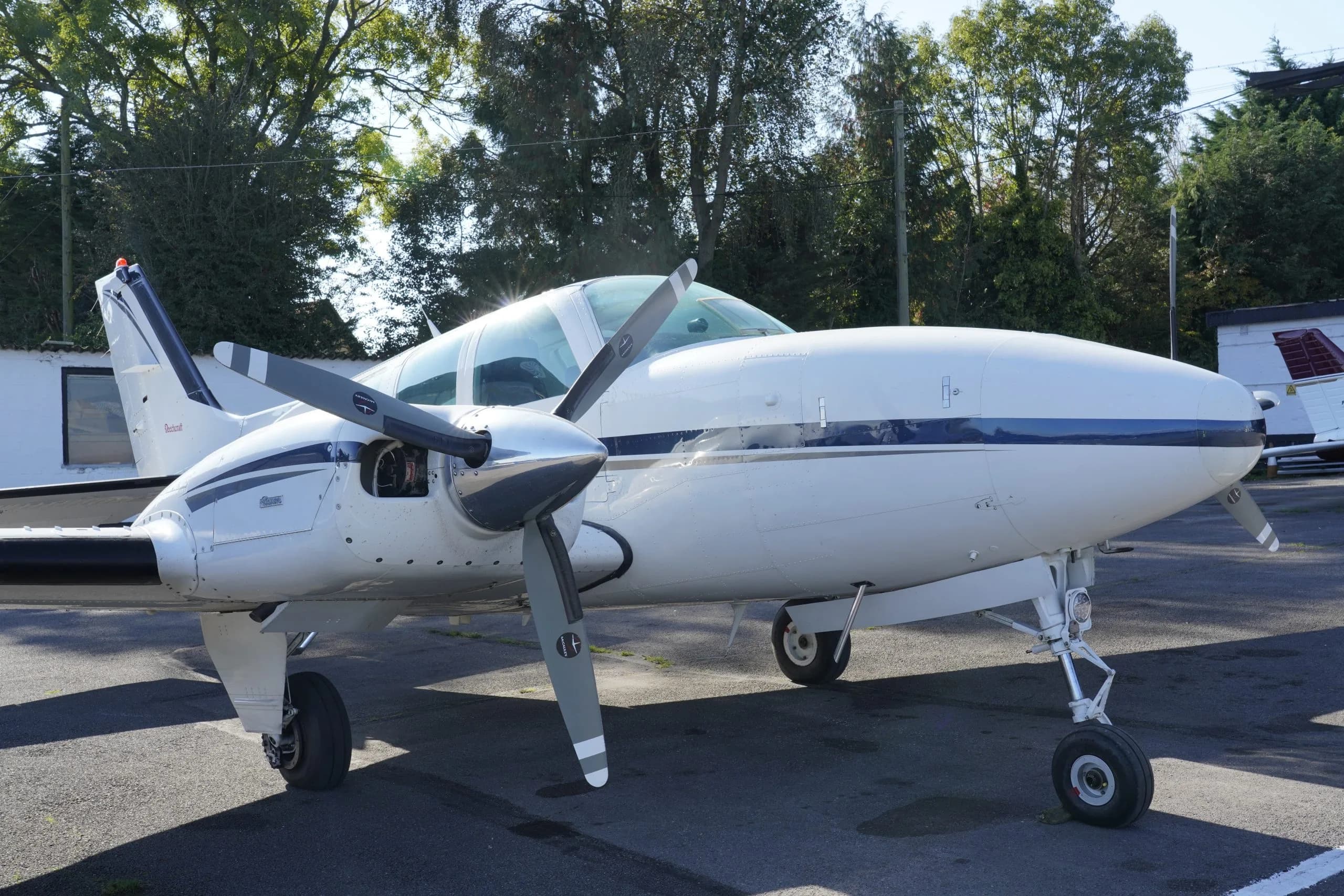 A front-side view of a white twin-engine propeller aircraft with blue stripes, parked on an asphalt apron.