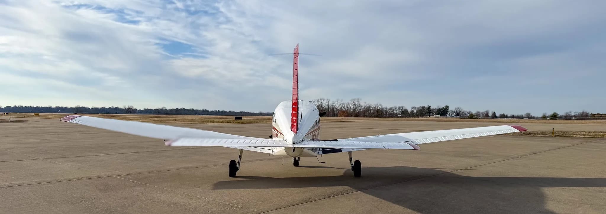 A rear view of a white and red Piper Archer TX aircraft parked on an airfield under a blue sky with scattered clouds.