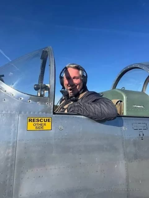A smiling pilot in a headset looks out from the cockpit of a silver experimental aircraft under a clear blue sky.