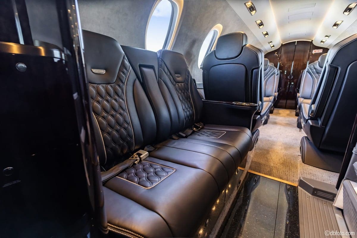 A close-up view of luxurious black leather aircraft cabin seats with quilted stitching and gold accents, featuring a side table and large oval windows.