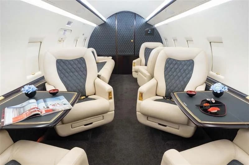 A luxurious private jet cabin with cream and dark grey quilted seats and tables set with sushi and a magazine.