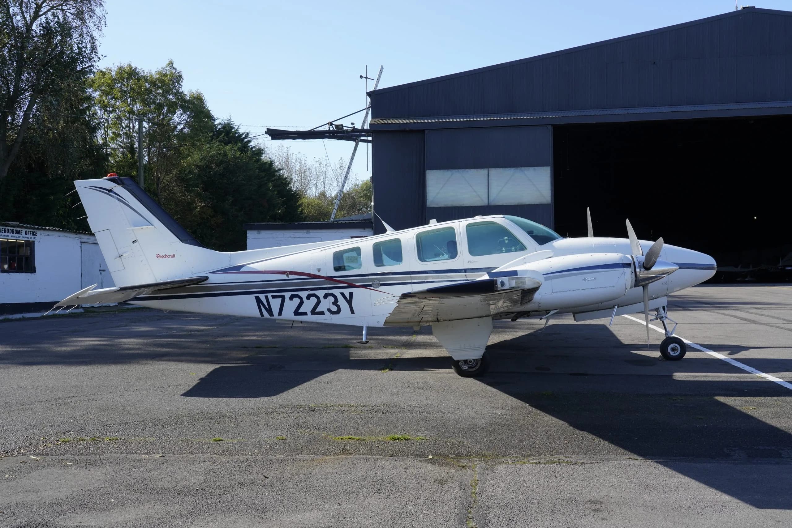 A white twin-engine propeller aircraft with blue and red stripes, registration N7223Y, parked on an asphalt apron next to a hangar.