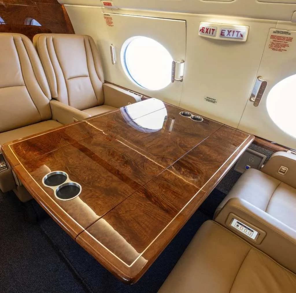 A close-up view of a large polished wood table with cup holders and tan leather seats in the aircraft cabin.