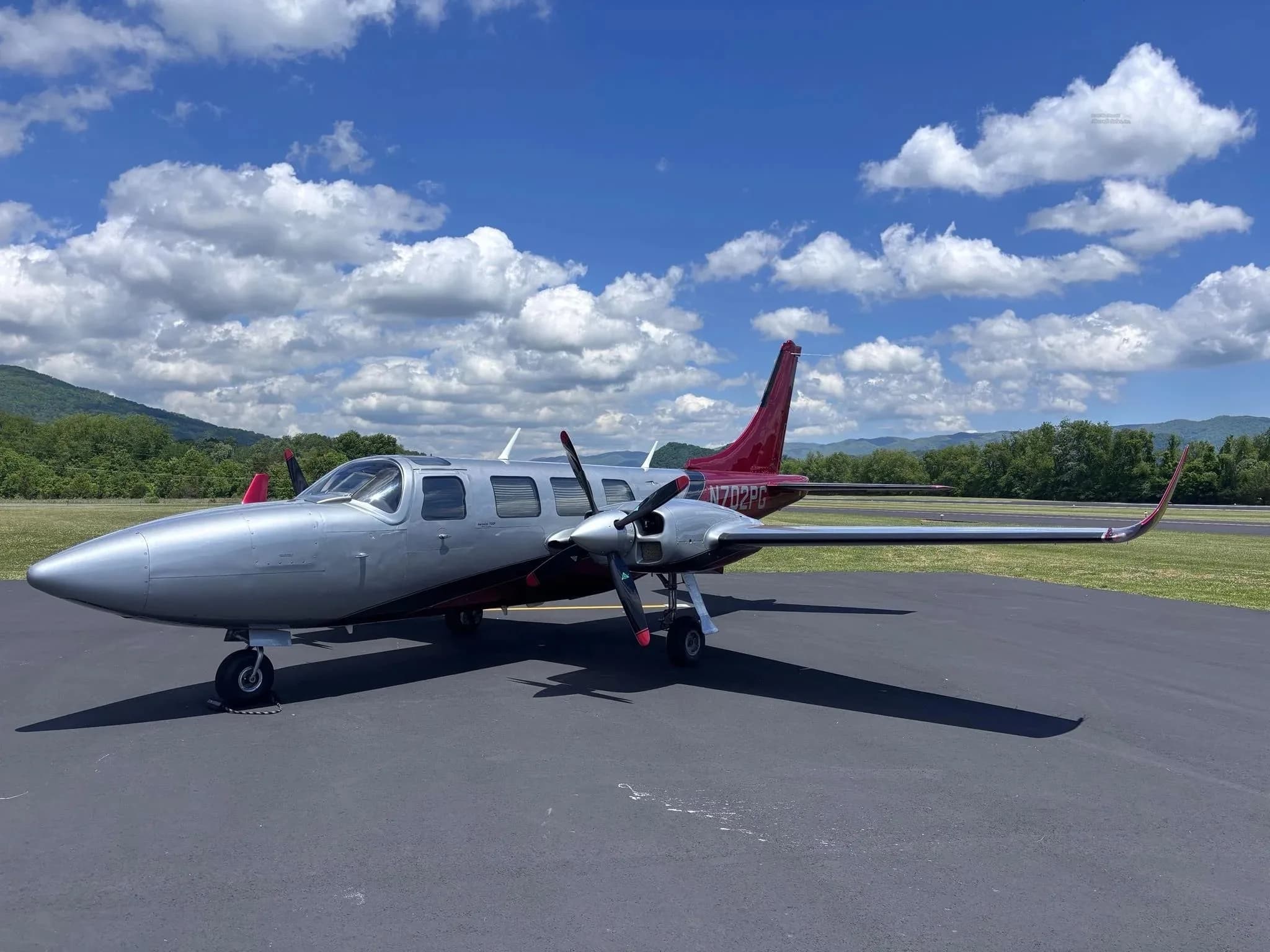 A silver and red twin-engine Aerostar aircraft (N702PG) parked on an airfield with mountains in the background.