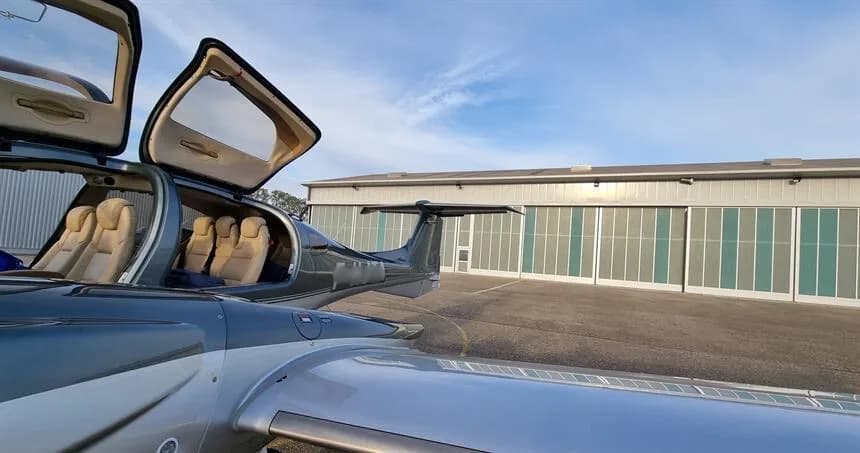 The open doors of a dark blue and silver Diamond DA62 aircraft reveal its light-colored cabin seats on an airport apron.
