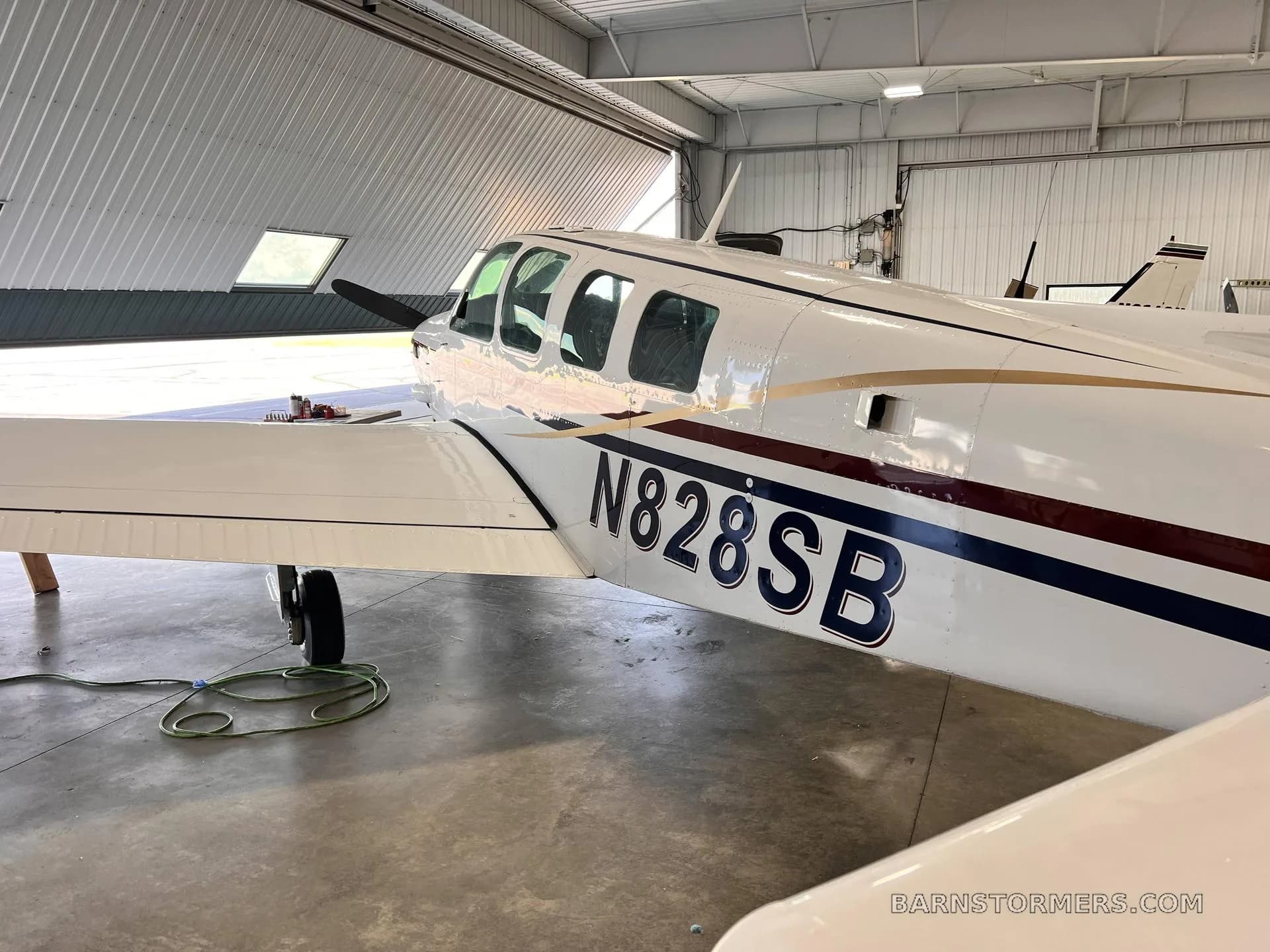 A white single-engine aircraft with dark blue and gold stripes and tail number N828SB is parked inside a hangar.