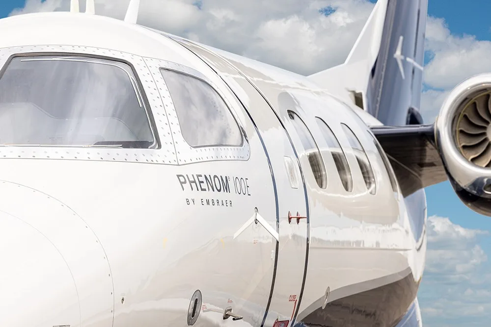 A close-up view of the white fuselage of the Embraer Phenom 100E, showing the cockpit windows and passenger door.