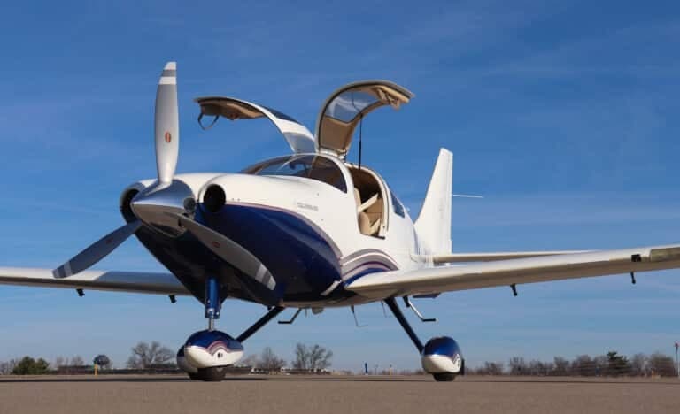 Front view of a white and blue single-engine propeller aircraft with gull-wing doors open on an airfield.
