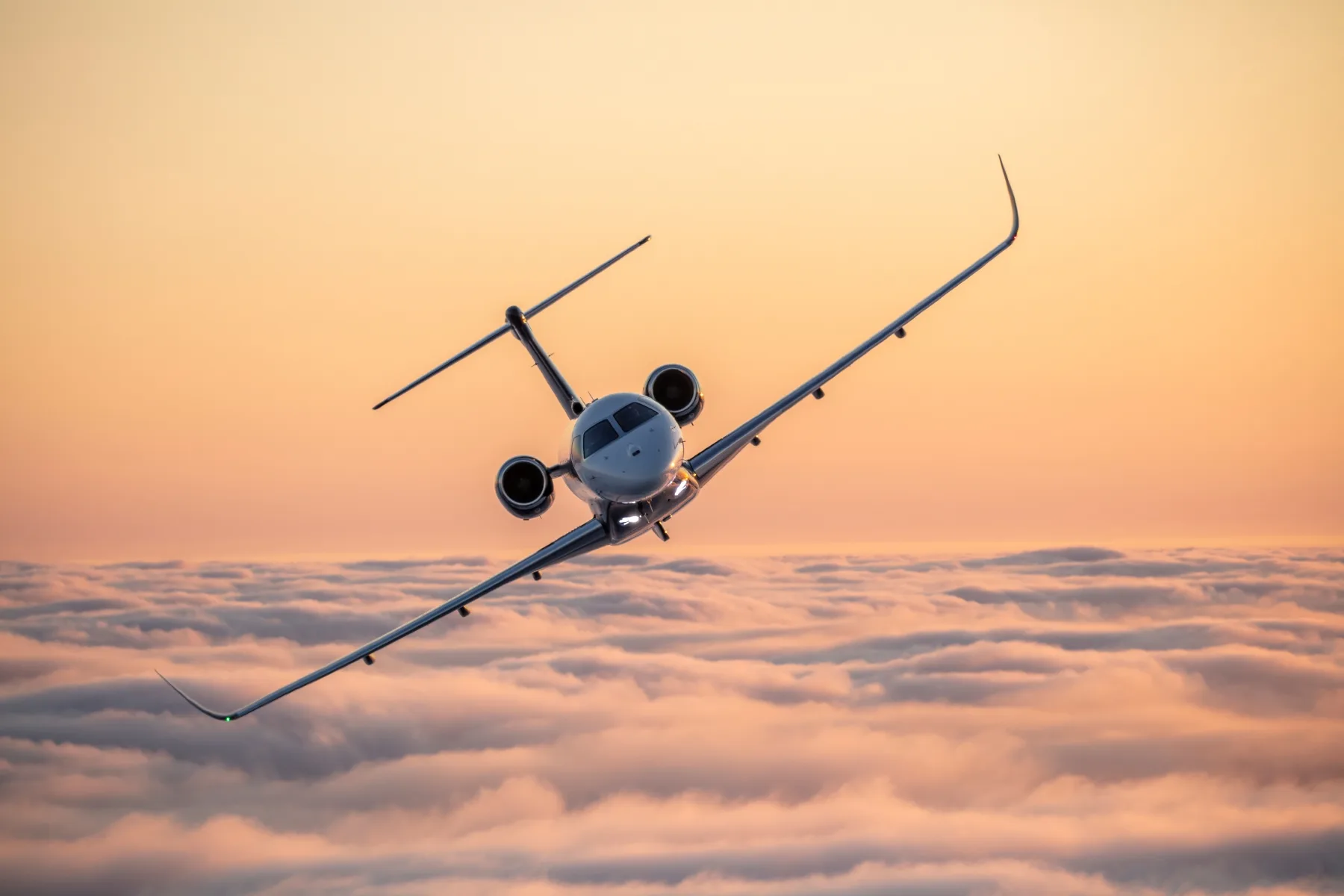 A white business jet with dark engines flies head-on over clouds during sunset.