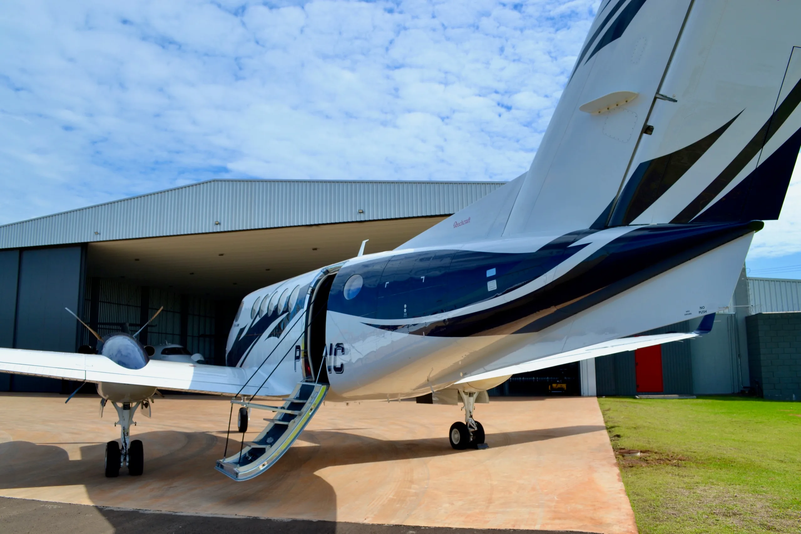 A rear-side view of a white and blue twin-turboprop aircraft with its passenger door open and stairs extended, next to a hangar.