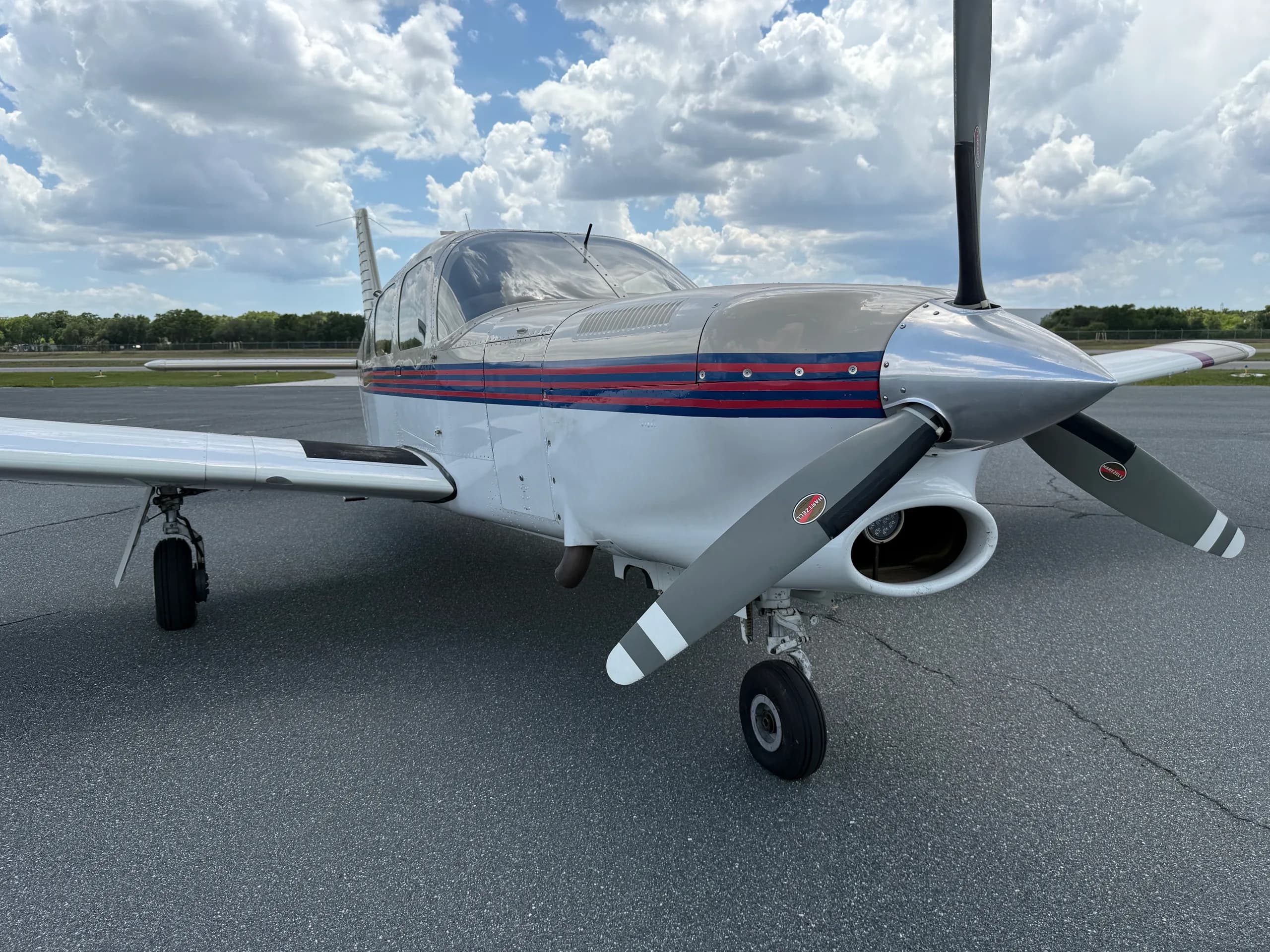 A white and grey single-engine propeller aircraft with red and blue stripes is parked on an asphalt tarmac under a cloudy sky.