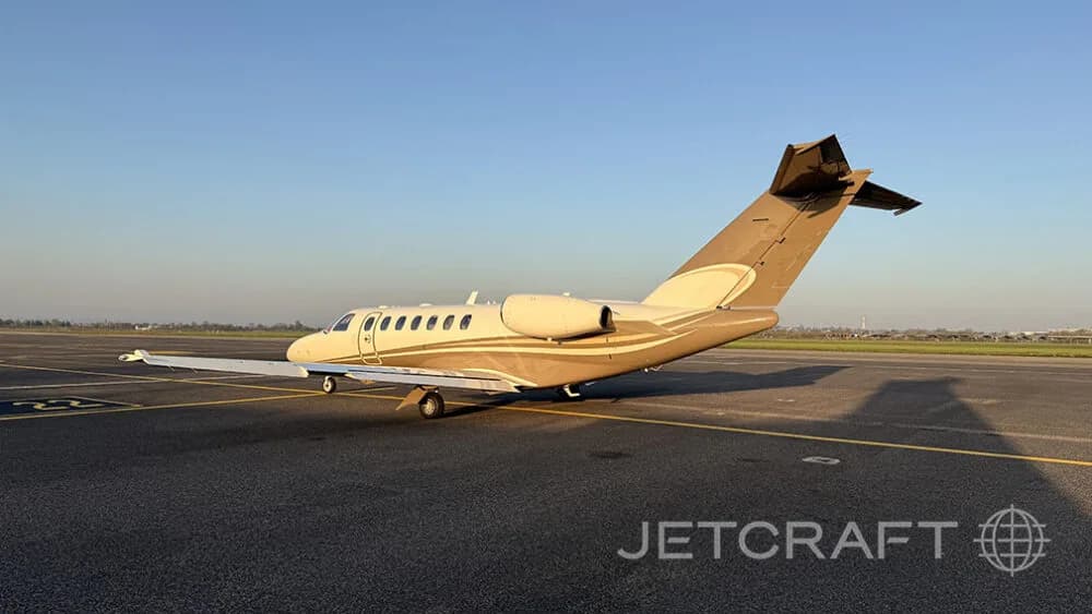 A beige and brown Cessna Citation business jet parked on an airport tarmac from a rear-side view.