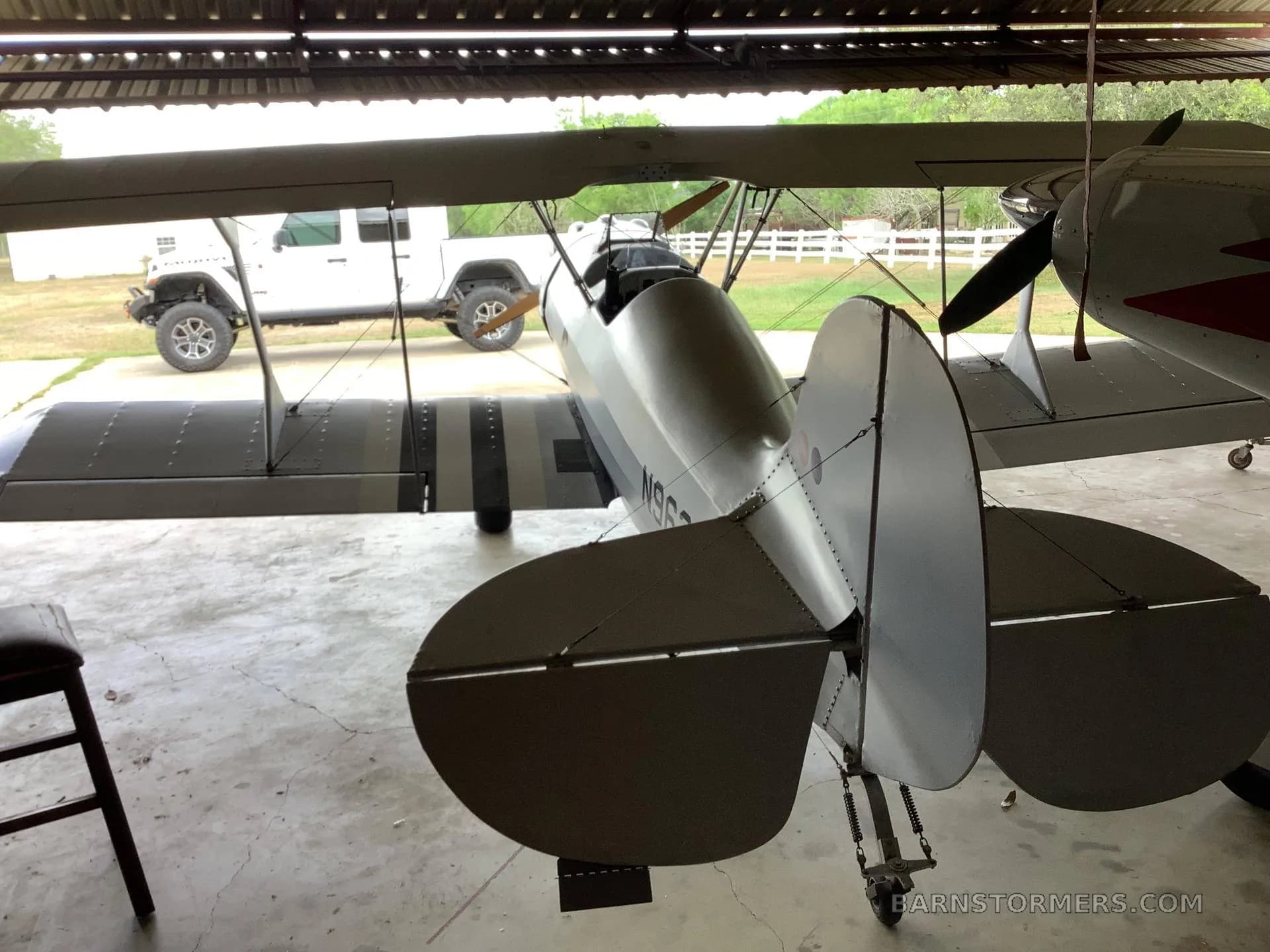 The tail and rear fuselage of a silver biplane are visible inside a hangar, with a white Jeep parked outside.
