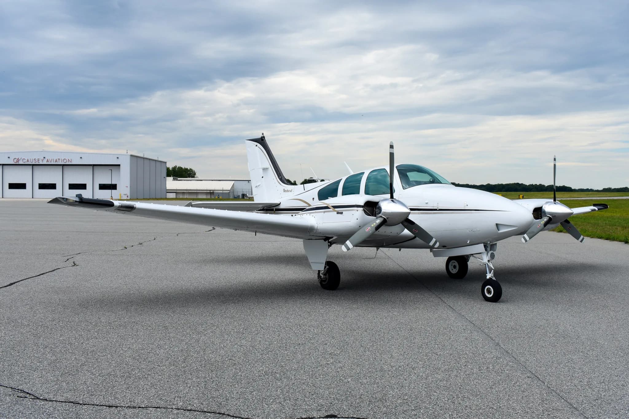 A white and black Beechcraft Baron aircraft, N6747V, is parked on an asphalt runway with hangars in the background.