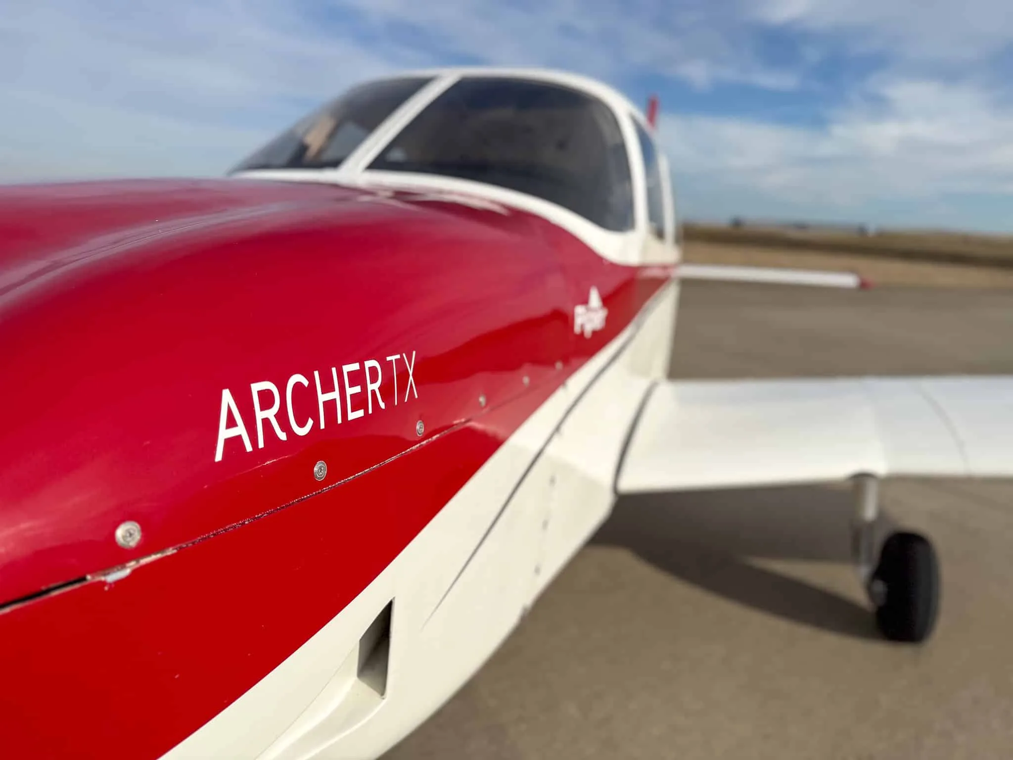 A close-up of the red and white fuselage of a Piper Archer TX aircraft, showing the "ARCHER TX" text.