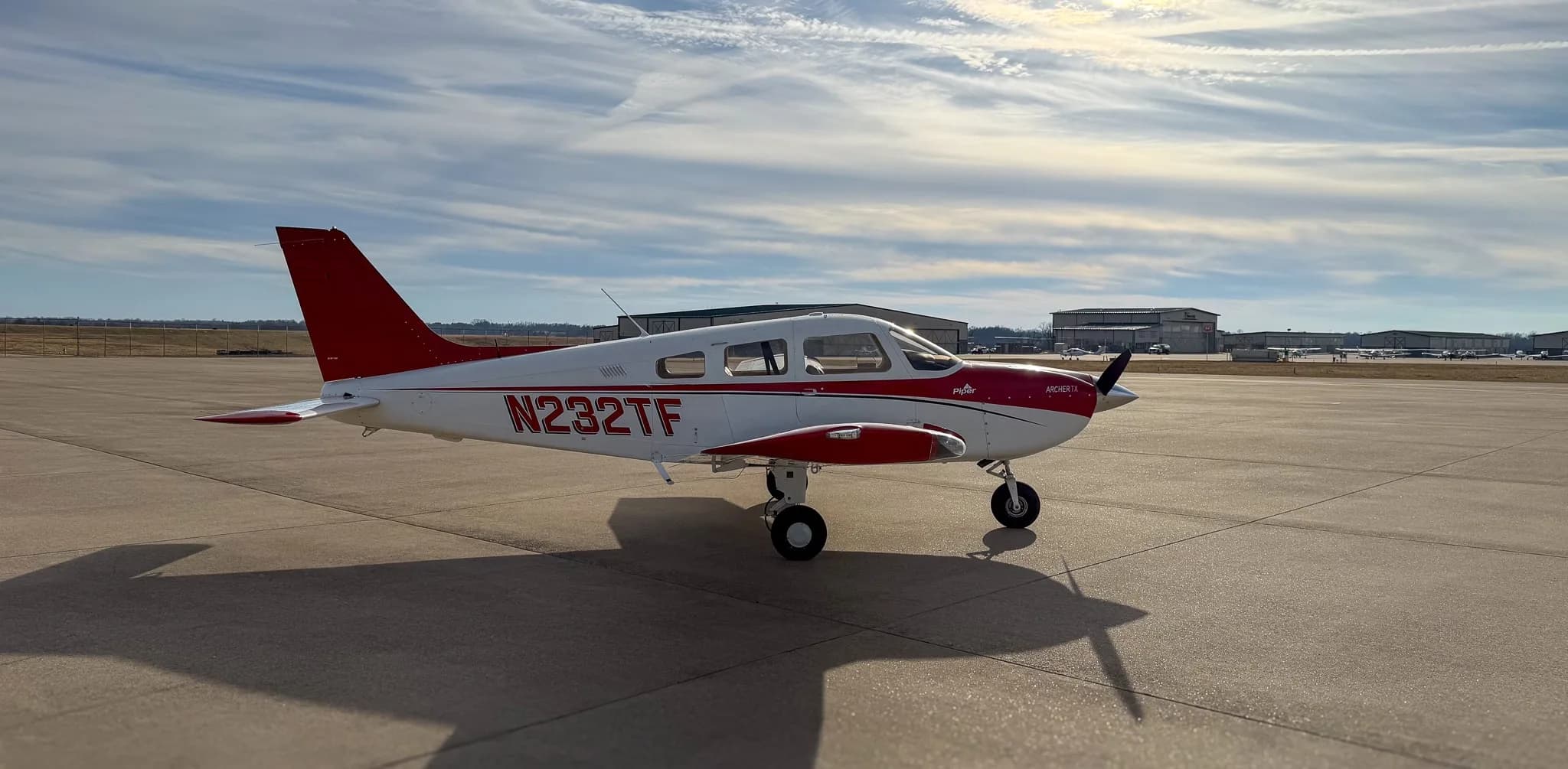 A three-quarter front-right view of a white and red Piper Archer TX, N232TF, parked on an airfield with hangars in the background.