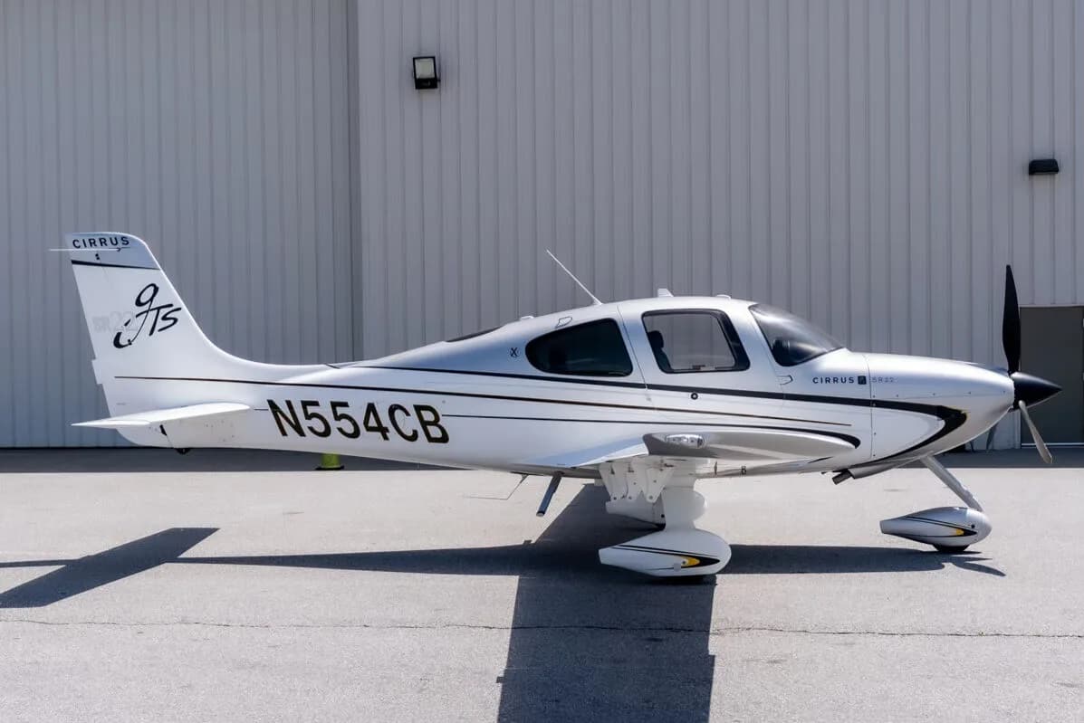 A side profile view of a silver and white Cirrus SR22T aircraft with registration N554CB parked in front of a hangar.