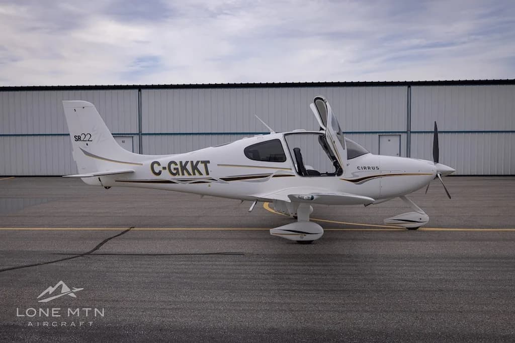 A side view of a white Cirrus SR22 aircraft with registration C-GKKT, doors open, parked on an airfield.